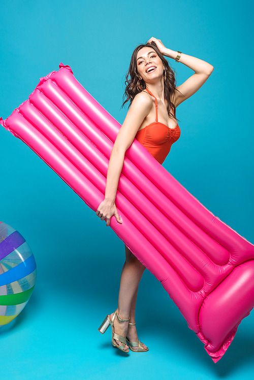 full length view of smiling woman in swimsuit holding inflatable swimming mattress on blue
