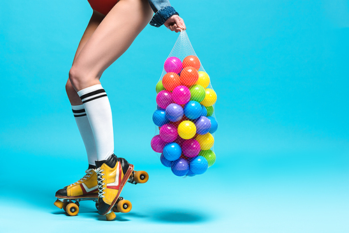 cropped view of woman in knee socks and roller skates holding string bag with colorful balls on blue