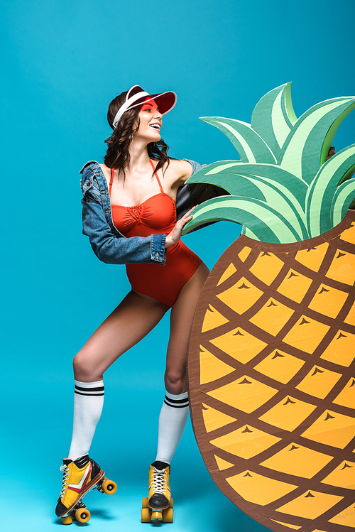full length view of woman in swimsuit and roller skates standing near decorative pineapple on blue