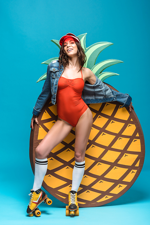 full length view of woman in swimsuit and roller skates standing near decorative pineapple on blue