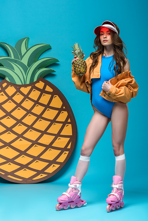 full length view of girl in swimsuit and roller skates holding pineapple on blue