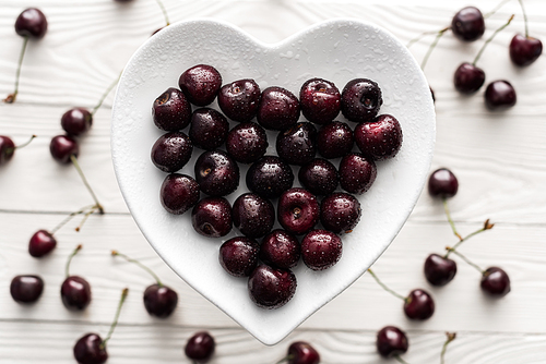 top view of fresh, sweet and ripe cherries covered with water drops on heart shaped plate