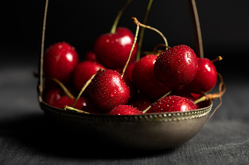 selective focus of wet red delicious cherries in metal basket on wooden dark table isolated on black