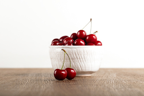 selective focus of red delicious cherries in white bowl and on wooden table isolated on white