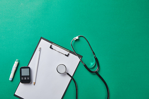 top view of blood lancet and stethoscope near glucometer and pencil on folder on green background