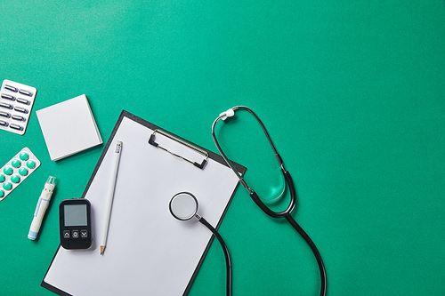 top view of blood lancet, sticky notes, blister packs and stethoscope near glucometer and pencil on folder on green background