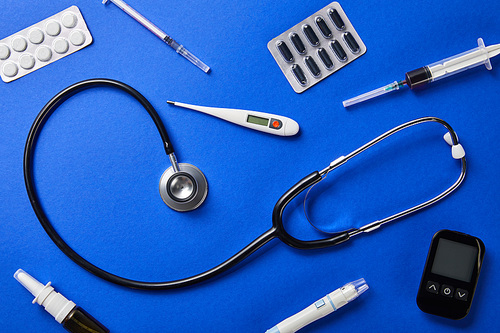blister packs, nasal spray, thermometer, blood lancet, glucometer, syringes and stethoscope on blue background