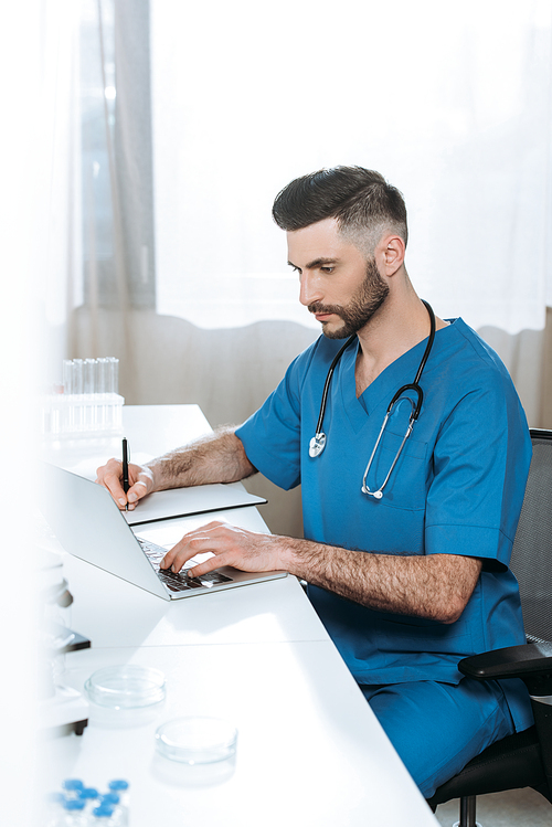 selective focus of attentive doctor writing while typing on laptop
