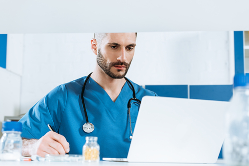 selective focus of young doctor writing while looking at laptop