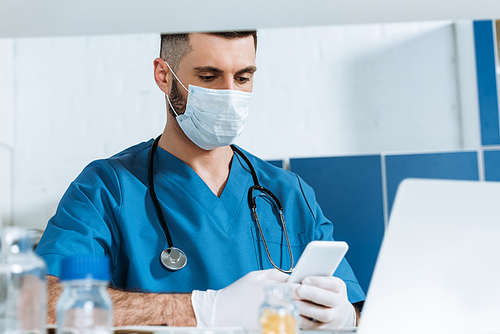 young doctor in medical mask and latex gloves using smartphone near laptop