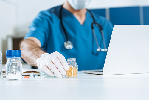 cropped view of doctor in latex gloves taking glass container with capsules while sitting near laptop