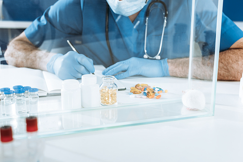 cropped view of veterinarian in medical mask and lates gloves writing near containers with medicines and white mouse in glass box