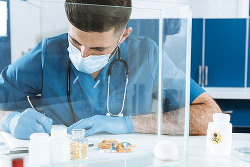 young veterinarian in medical mask and lates gloves writing near containers with medicines and white mouse in glass box