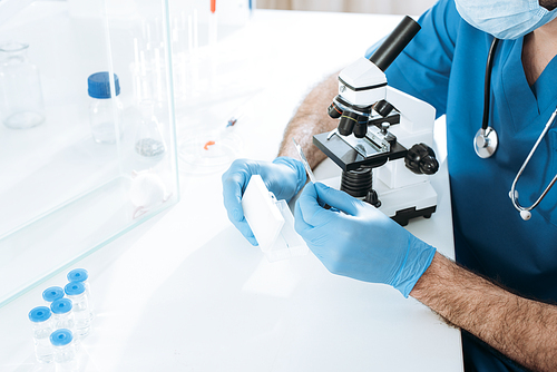 cropped view of biologist in medical mask and lates gloves holding test box while making analysis with microscope near white mouse in glass box