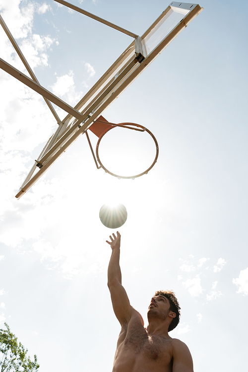bottom view of shirtless basketball player throwing ball in basket in sunny day