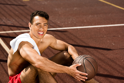 shirtless mixed race basketball player smiling while sitting at basketball court with ball in sunny day
