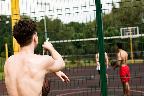 back view of shirtless basketball player with ball looking at friends and pointing with finger at basketball court