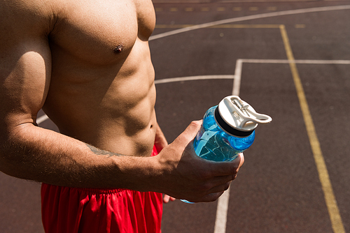partial view of shirtless muscular sportsman holding sport bottle at basketball court