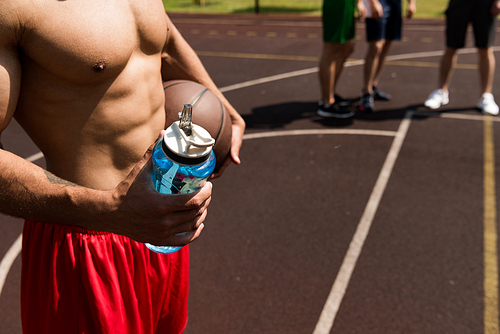 partial view of shirtless muscular sportsman holding sport bottle and ball at basketball court
