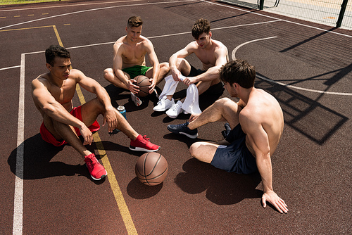 four tired shirtless basketball players sitting at basketball court in sunny day