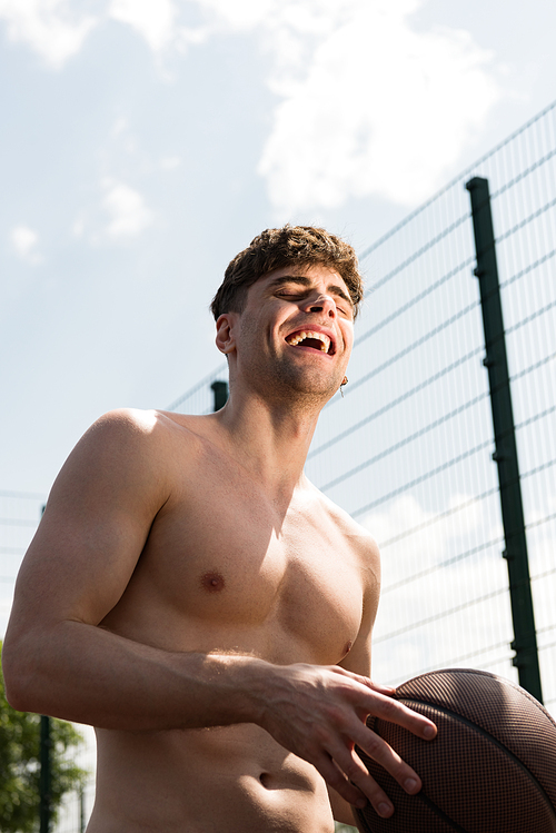 smiling sexy shirtless sportsman holding ball at basketball court under blue sky