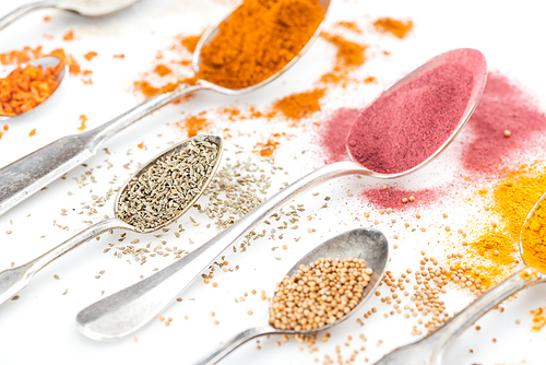 close up view of various bright spices in silver spoons on white background