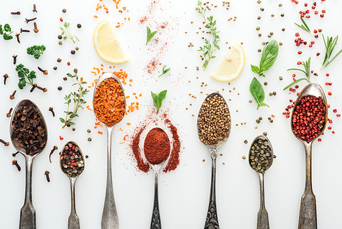 top view of colorful spices in silver spoons near herbs and lemon slices on white background