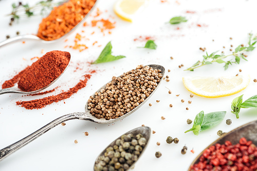 close up view of colorful spices in silver spoons near herbs and lemon slices on white background