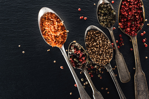 top view of pepper, candied fruit and mustard in silver spoons on black textured background