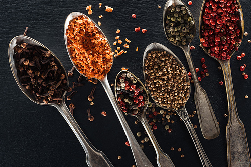 top view of pepper, candied fruit, coriander and carnation seeds in silver spoons on black textured background