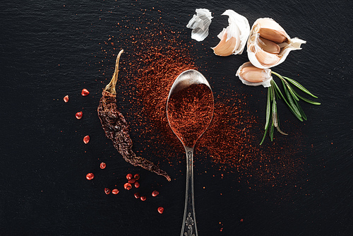 top view of red pepper powder in silver spoon on black background with dried chili peppe, herbs and garlic