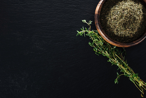 top view of thyme spice in wooden bowl near green herb on black background