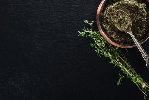 top view of dried thyme in wooden bowl with spoon near green herb on black background