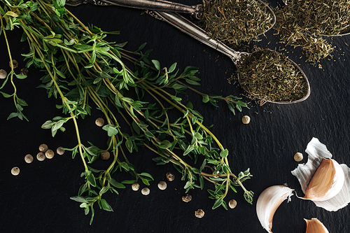 top view of dried thyme in silver spoons near green herb, white pepper and garlic cloves on black background