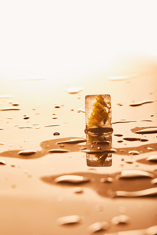 transparent ice cube with frozen flower near water drops on yellow illuminated background