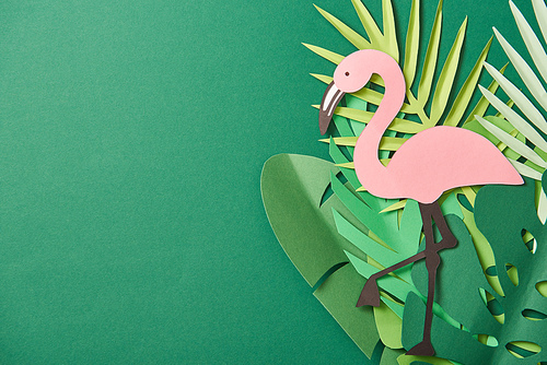 top view of paper cut palm leaves and pink flamingo on green background with copy space