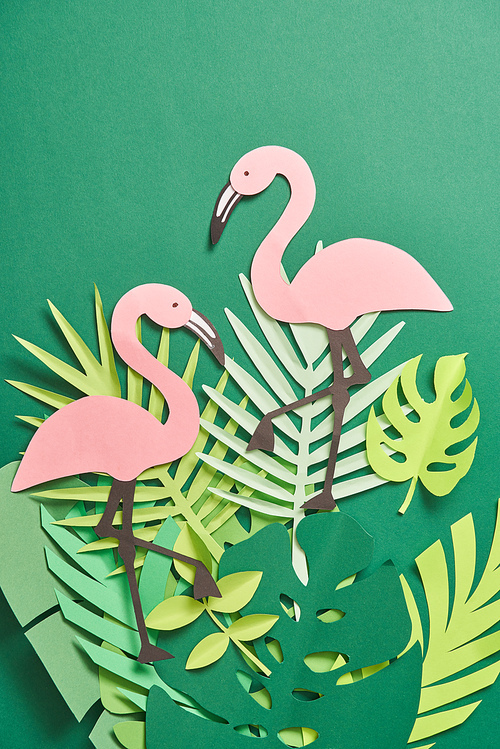 top view of exotic paper cut palm leaves and pink flamingos on green background with copy space