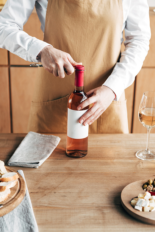 cropped view of sommelier in apron opening bottle of wine at table