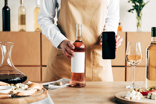 partial view of sommelier in apron standing near table with bottles of wine and showing smartphone with blank screen
