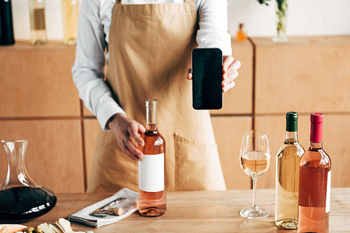 partial view of sommelier in apron standing near table with bottles of wine and showing smartphone with blank screen