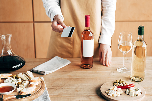 cropped view of sommelier in apron standing near table with bottles of wine and holding credit card
