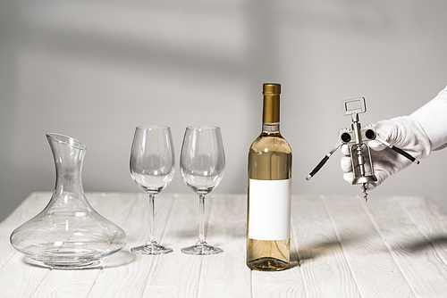 cropped view of waiter in white glove holding corkscrew near table with bottle of wine, wine glasses and jug