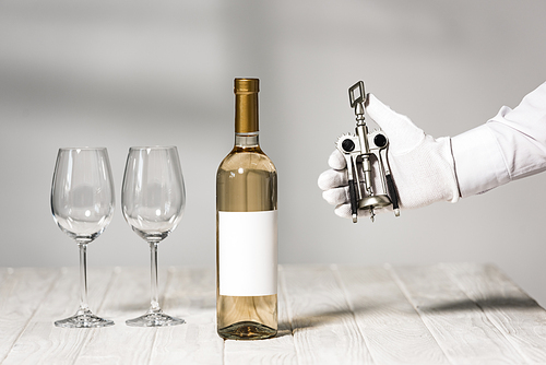cropped view of waiter in white glove holding corkscrew near table with bottle of wine and wine glasses