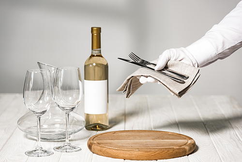 cropped view of waiter in white glove holding cutlery near table with bottle of wine, wine glasses and jug