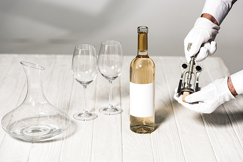 partial view of waiter in white gloves holding corkscrew on table near bottle, wine glasses and jug