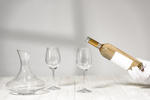cropped view of water in white glove holding bottle of wine near wine glasses on wooden table