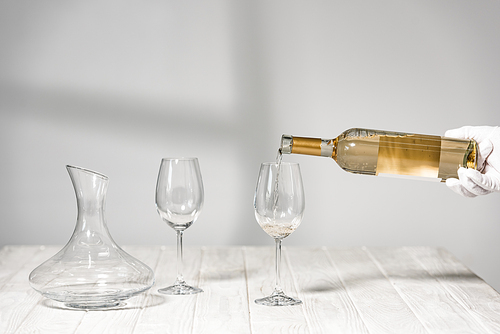 cropped view of water in white glove holding bottle of wine near wine glasses on wooden table