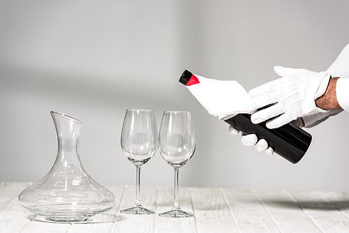 cropped view of water in white gloves holding bottle of wine near wine glasses on wooden table
