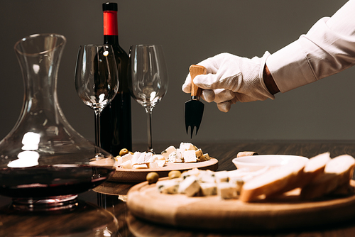 cropped view of waiter in white glove holding cheese fork near table with food and wine