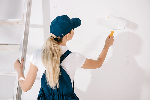 back view of young painter in uniform standing near white wall and holding paint roller